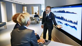 Europäischer Immobilienmakler im Gespräch mit Kunden in einem modernen deutschen Büro, gemeinsame Betrachtung eines Bildschirms mit Marktdaten