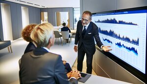 Europäischer Immobilienmakler im Gespräch mit Kunden in einem modernen deutschen Büro, gemeinsame Betrachtung eines Bildschirms mit Marktdaten